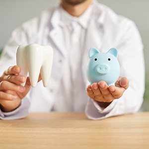  Shoulders-down view of dentist in white coat holding a model tooth and blue piggy bank