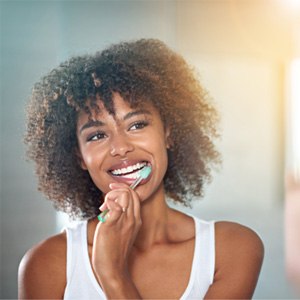Woman in white tank top brushing her teeth at a mirror