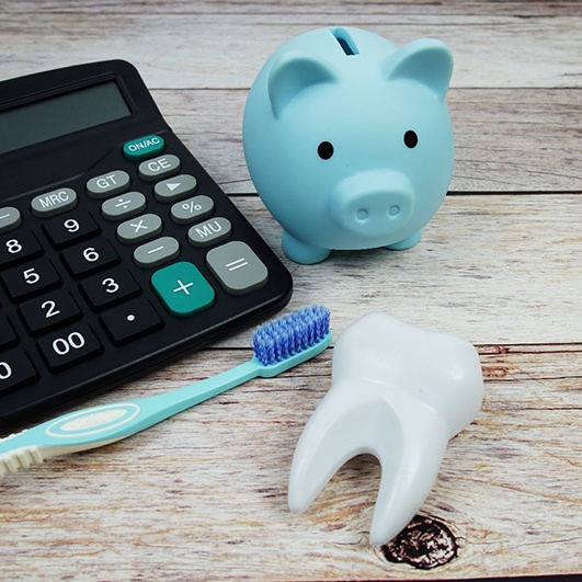 Blue piggy bank and toothbrush next to calculator and model tooth on wooden surface